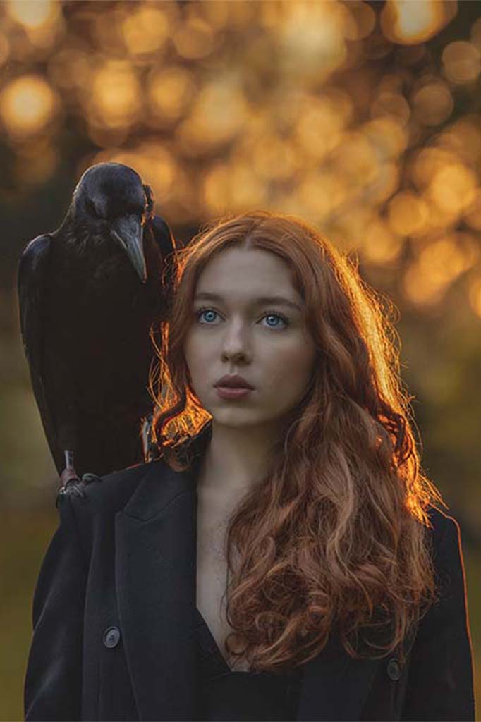 Portret kobiety w czarnym płaszczu z rudymi włosami o zachodzie słońca, na jej ramieniu siedzi czarny kruk.