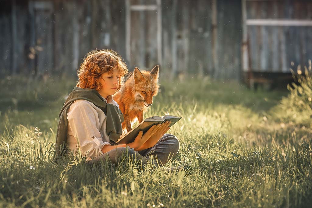 Chłopiec siedzący na trawie i czytający książkę, zza jego ramienia wychyla się lis.