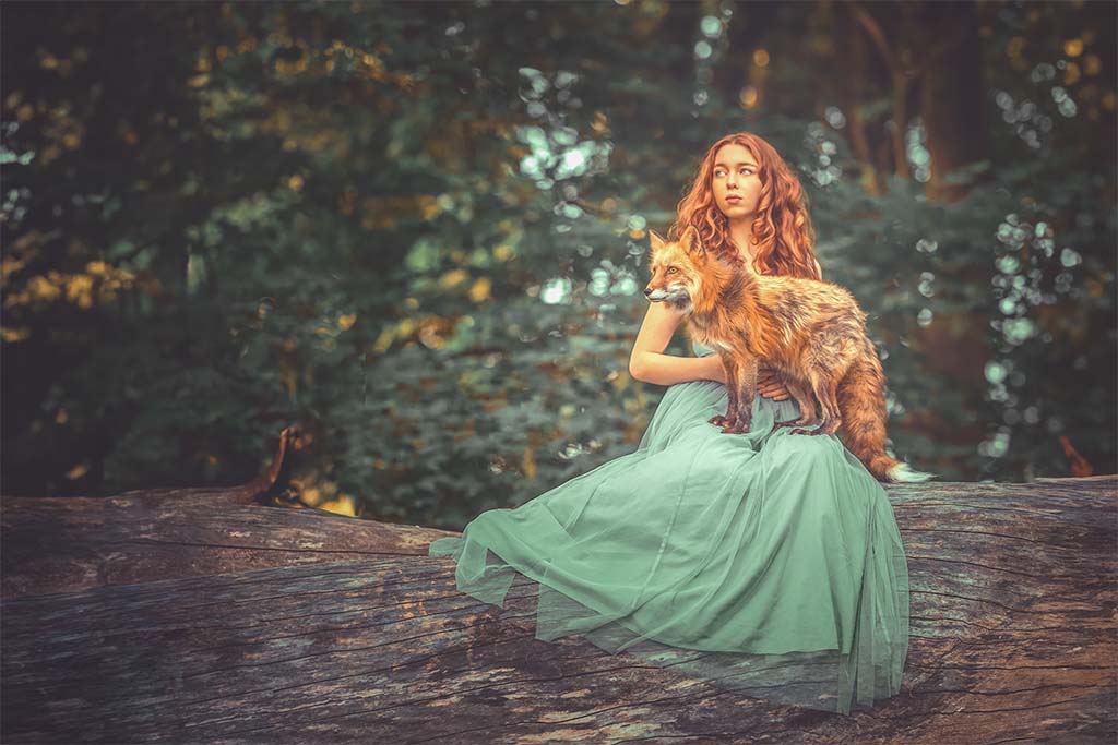 Kobieta w zielonej tiulowej sukience z rudymi włosami siedząca na konarze drzew. Na jej kolanach lis.