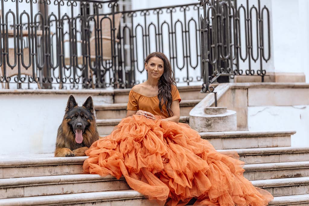 Pałacowe schody, na nich siedząca kobieta w pomarańczowej tiulowej sukni, obok niej leży pies.