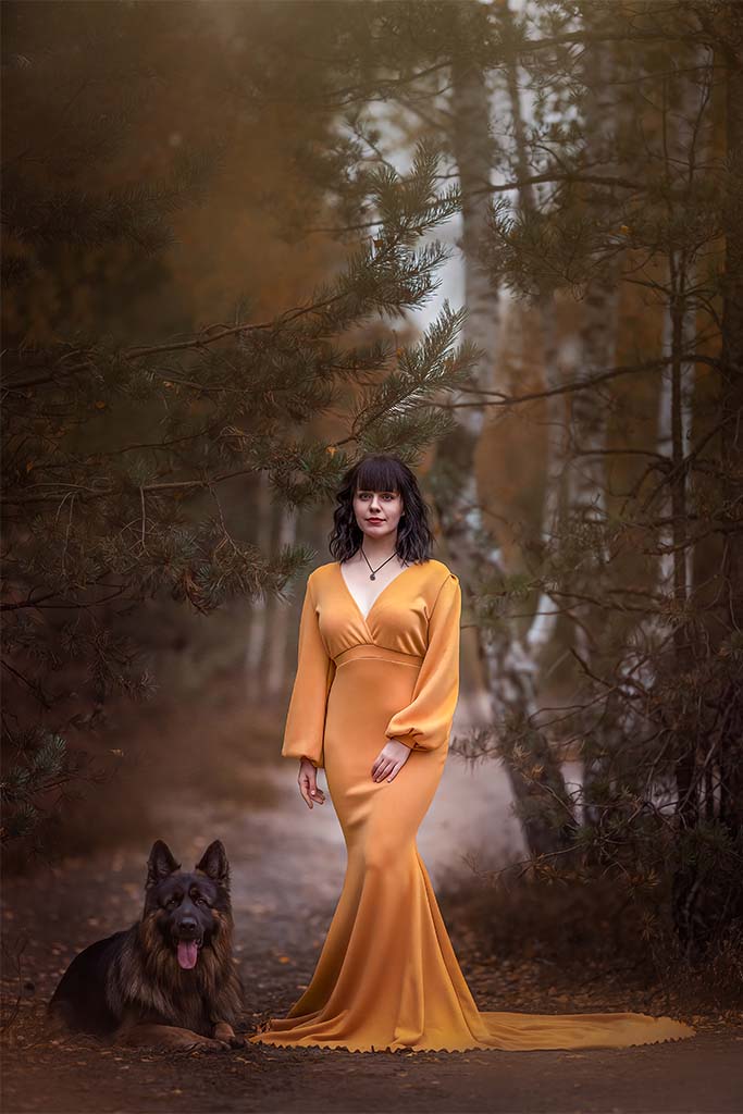 Kobieta stojąca w lesie pomiędzy drzewami w pomarańczowej sukni, obok niej leży pies.