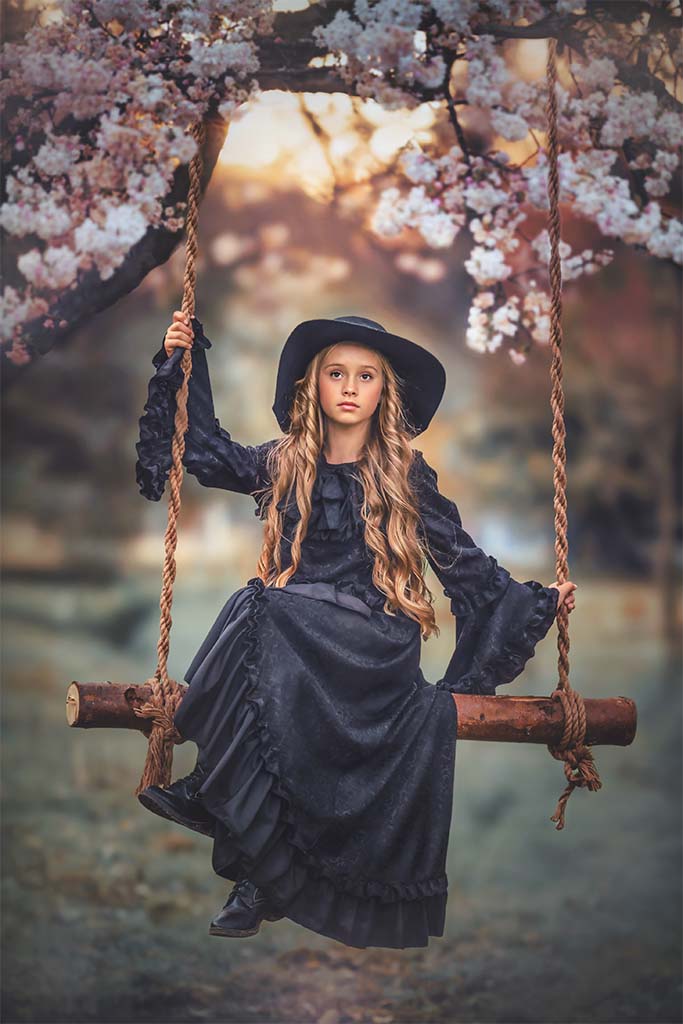 dziewczynka w czarnej sukience i czarnym kapeluszu na drewnianej huśtawce na łące