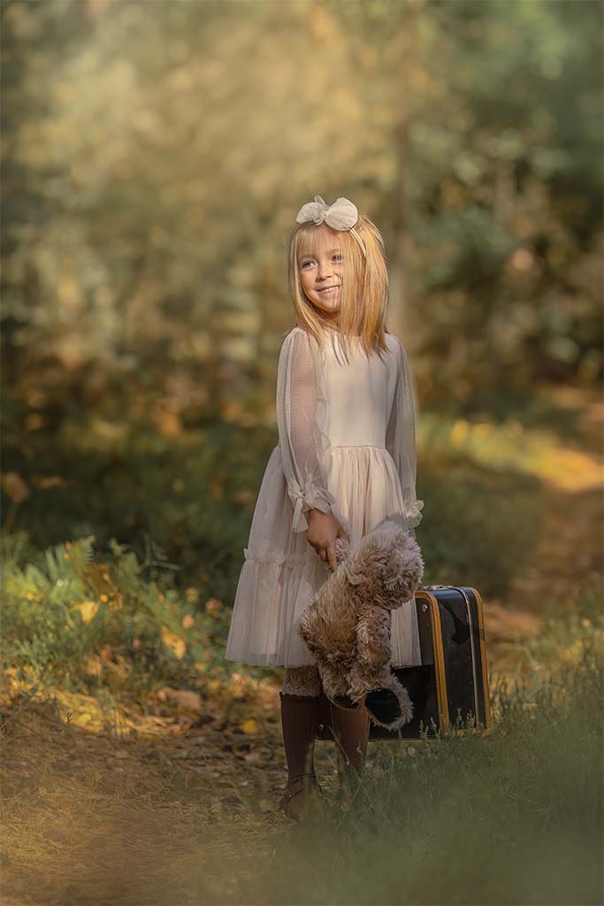 dziewczynka w białej sukience trzymająca w rękach misia i walizkę na leśnej drodze