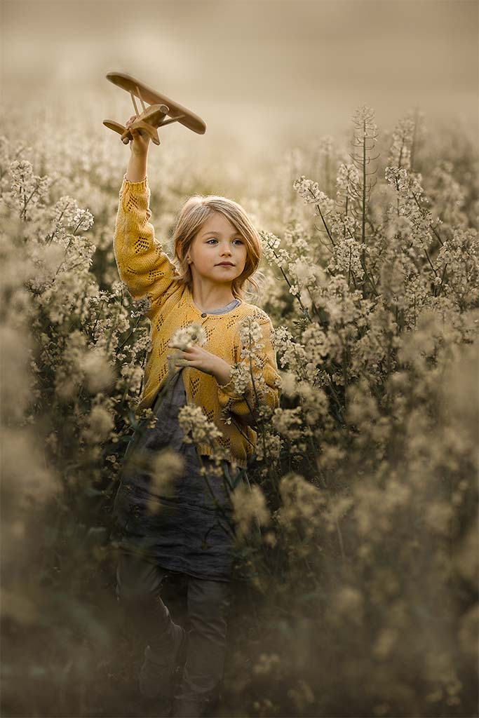 dziewczynka w żółtym sweterku z drewnianym samolotem w ręku w polu rzepaku