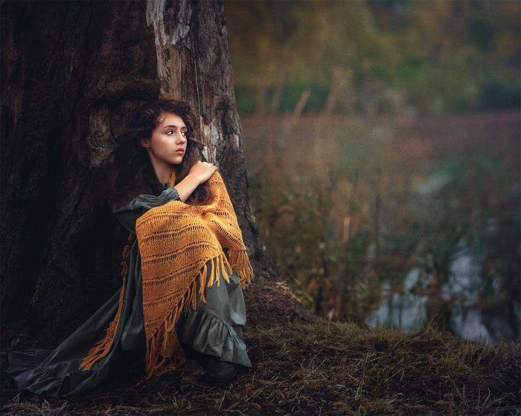 dziewczynka siedząca pod drzewem w z pomarańczową chustą na ramionach patrząca w dal