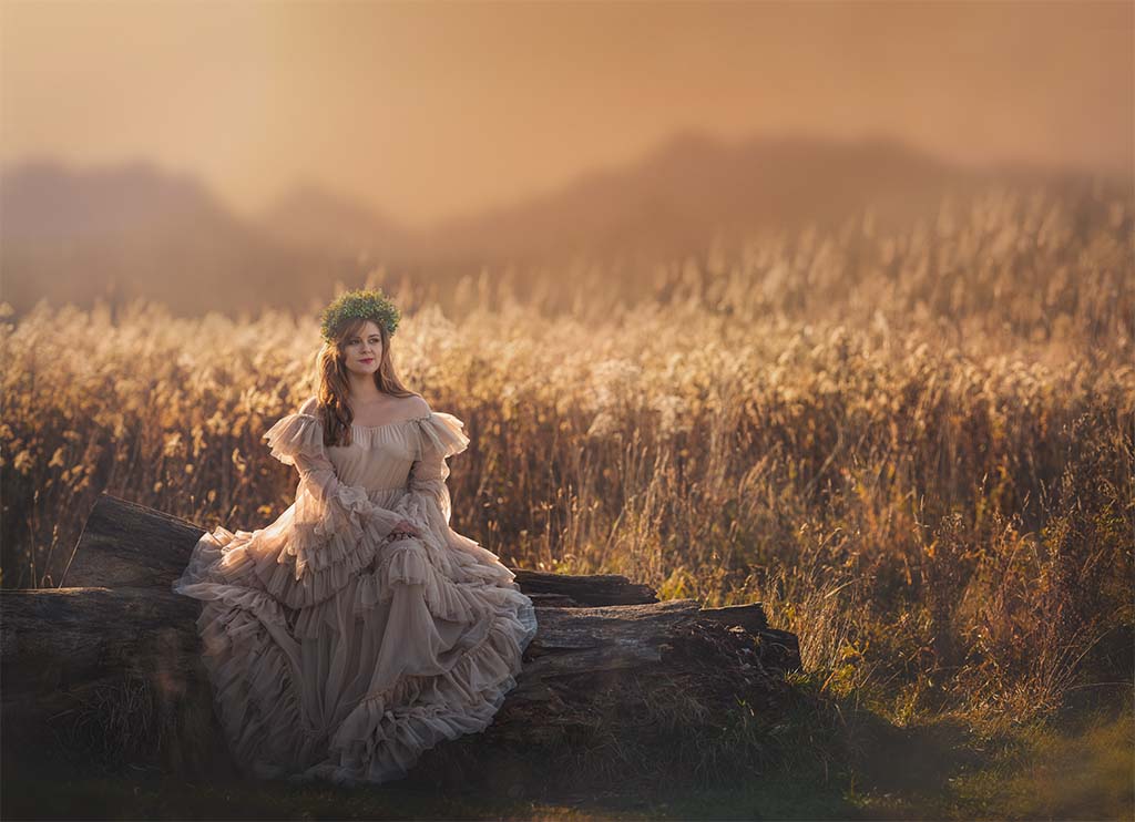 Kobieta w kremowej tiulowej sukni i zielonym wianku na głowie siedząca na konarze drzewa