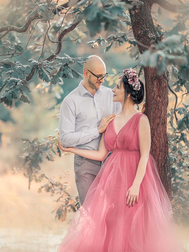 kobieta w różowej sukni stojąca pod drzewem oparta o mężczyznę