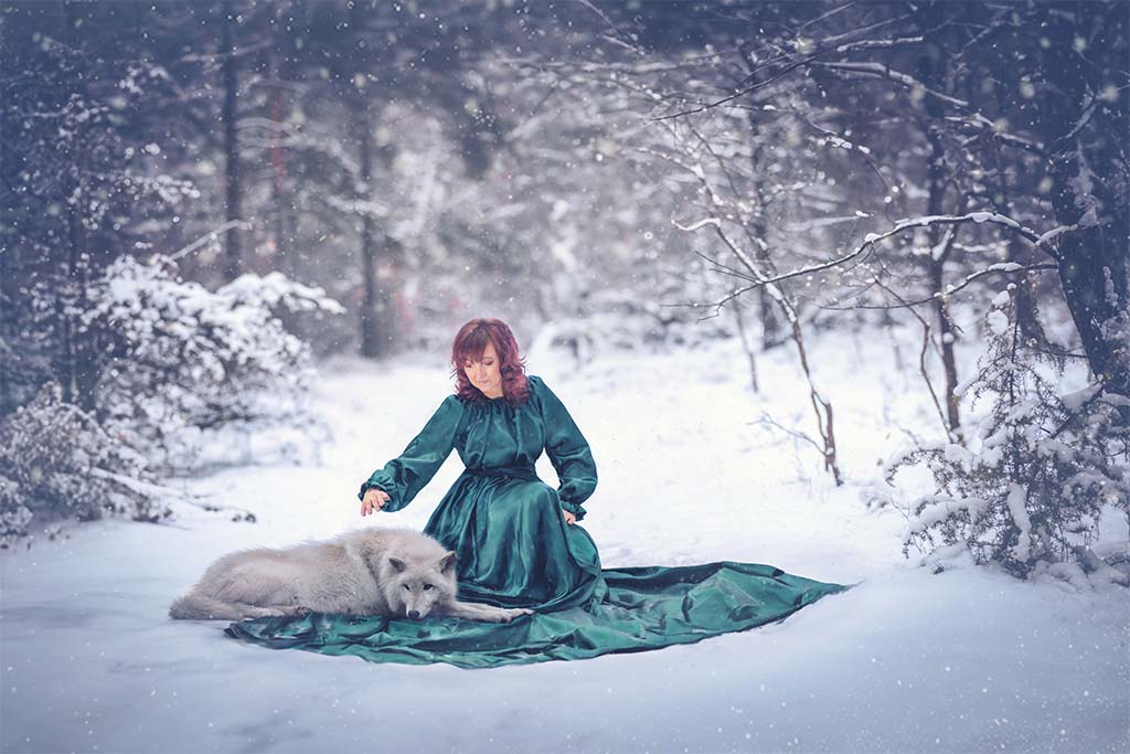 Zimowy las, na śniegu siedząca kobieta w zielonej atłasowej sukience, na jej sukience leży biały wilk.