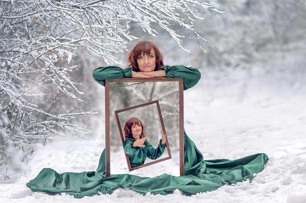 zimowy las, kobieta siedząca na śniegu oparta o ramę ze zdjęciem samej siebie
