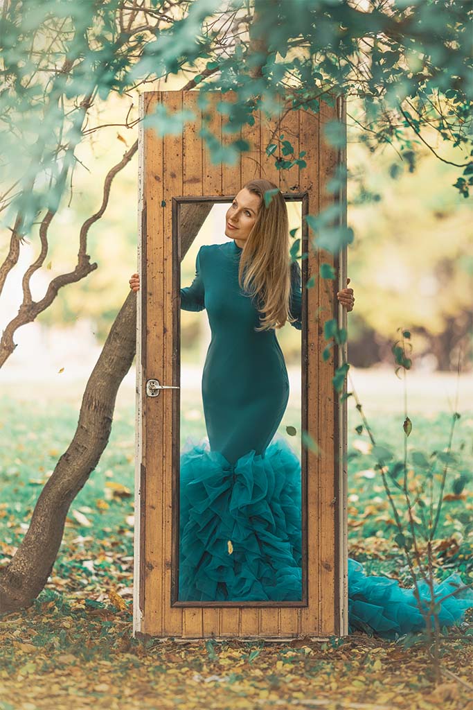 kobieta w zielonej sukni pozuje pomiędzy drzewami oparta o stare drzwi