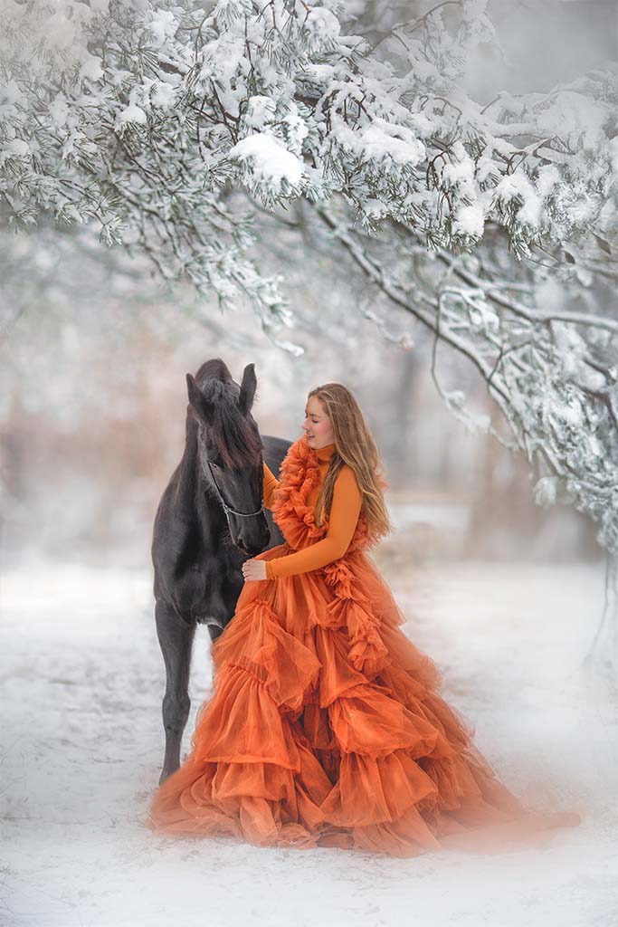 Kobieta w pomarańczowej tiulowej sukience pozująca z czarnym koniem na zimowej drodze.