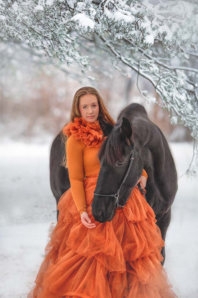 Kobieta w pomarańczowej tiulowej sukience pozująca z czarnym koniem na zimowej drodze.