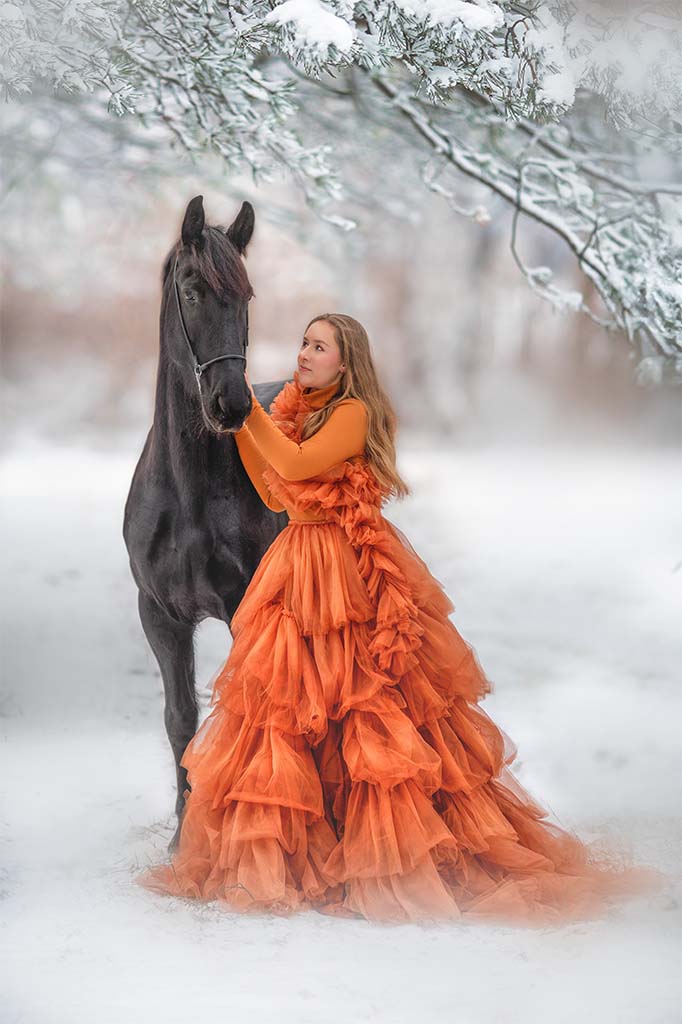 Kobieta w pomarańczowej tiulowej sukience pozująca z czarnym koniem na zimowej drodze.