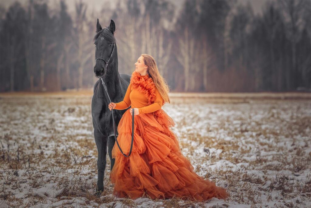 Kobieta w pomarańczowej tiulowej sukience pozująca z czarnym koniem na zimowej łące.