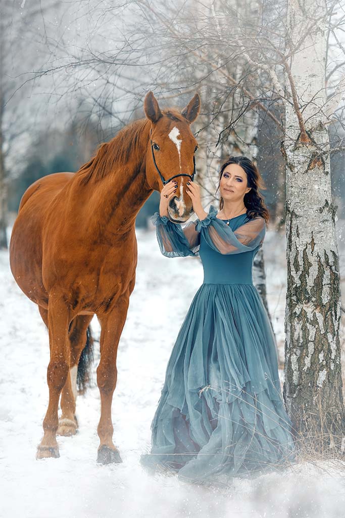 Młoda kobieta stojąca oparta o brzozę z rudym koniem obok. Kobieta ubrana w niebieską tiulową sukienkę. W tle śnieg.