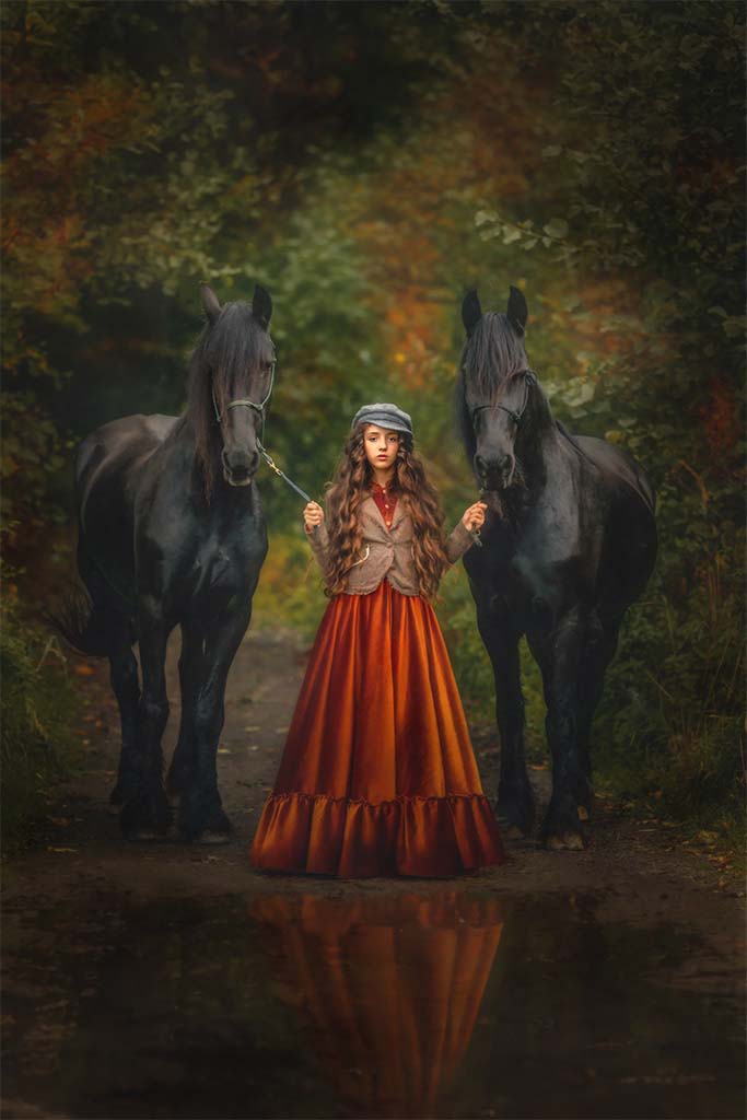 Młoda dziewczynka stojąca na drodze leśnej, trzymająca w rękach dwa czarne konie rasy fryzyjskiej