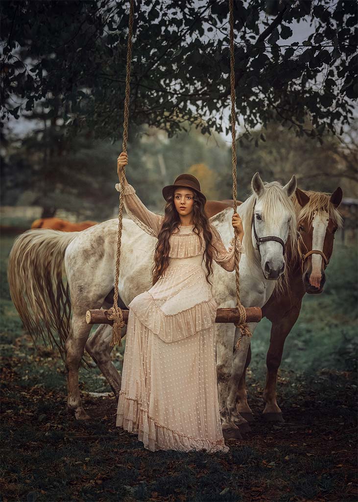 Młoda dziewczynka w kremowej sukience i brązowym kapeluszu siedząca na drewnianej huśtawce zamocowanej do drzewa, obok dwa konie